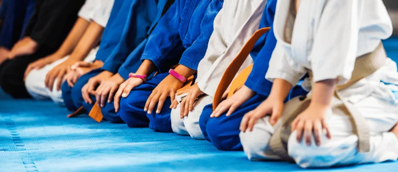 A row of martial arts students in white and blue gis with coloured belts, kneeling on blue mats at the start of a class.