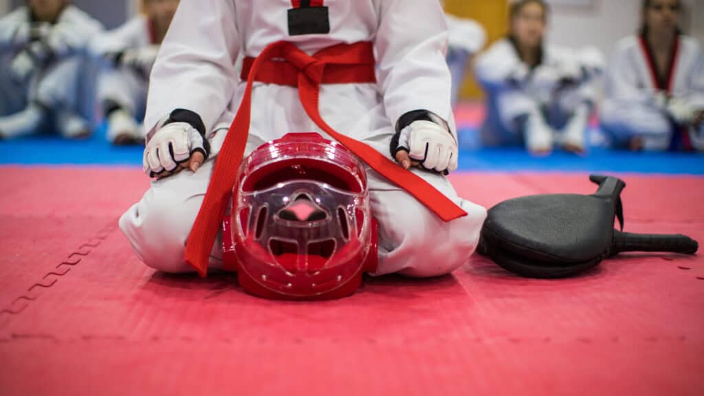 Martial artist in a red belt seated on a mat with protective gear and training equipment after joining a new dojo in January 2025.
