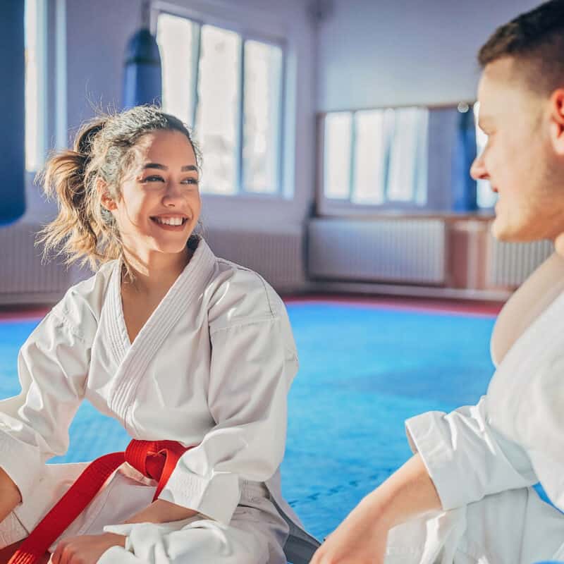 Two friends sit waiting for their karate class to begin after booking a spot on the mat next to each other.