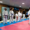 A group of people wearing martial arts uniforms stand in line on a mat, facing an instructor who is speaking to them.
