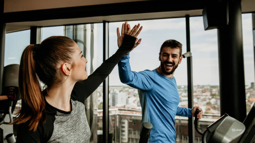 Two gym members smiling and high-fiving on cardio machines, highlighting positive member experience and community, a key factor when members choose a gym.